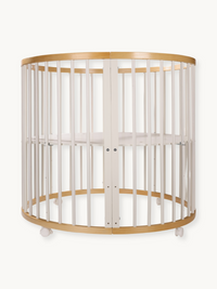 Round oval baby playpen with white vertical slats, natural wood top and bottom rails, lockable white wheels, front view on…
