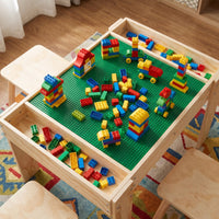 Overhead view of natural wood kids' activity table with green building plate covered in colourful stacked bricks, open sto…