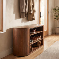 Walnut oval shoe bench with open shelves holding trainers and boots, styled in a sunlit entryway with coat hooks above