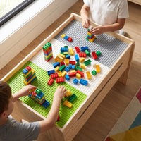 Two children playing with colourful building bricks on a natural wood table with green, white, and grey baseplate surface,…