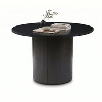 Black round dining table with ribbed pedestal base, styled with a dark vase, stacked books and a ceramic bowl, three-quart…