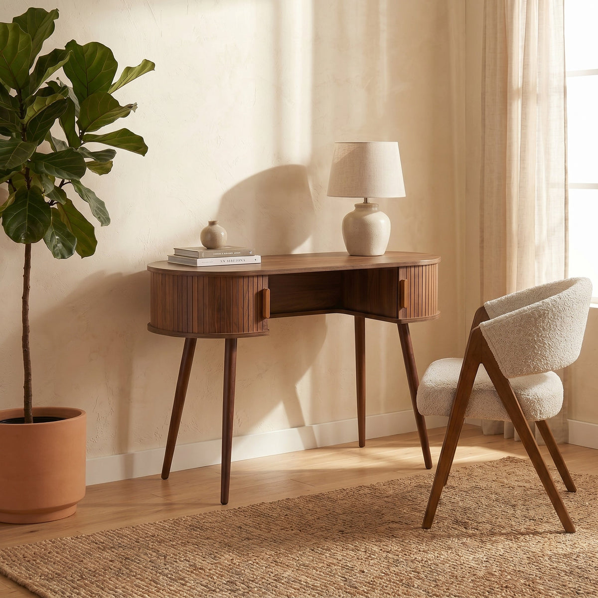 Walnut oval console table styled with a lamp, books, and boucle chair beside sheer curtains and a fiddle-leaf fig plant