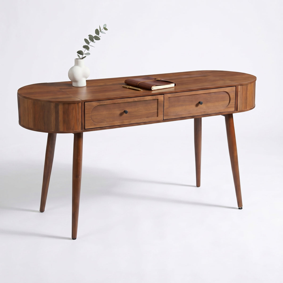 Walnut oval writing desk angled view, two drawers with black knobs, topped with white vase and leather notebook on white b…