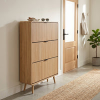 Tall oak-finish shoe cabinet styled in hallway entryway, beside open door, jute runner and fiddle-leaf fig plant nearby