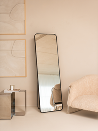 Black full-length standing mirror with rounded corners leaning against a beige wall beside a bouclé armchair, lifestyle se…