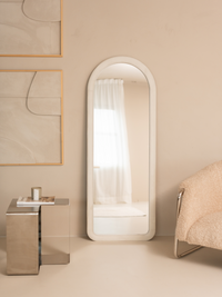 White suede velvet arched floor mirror leaning against beige wall, styled with chrome side table and boucle chair in neutr…