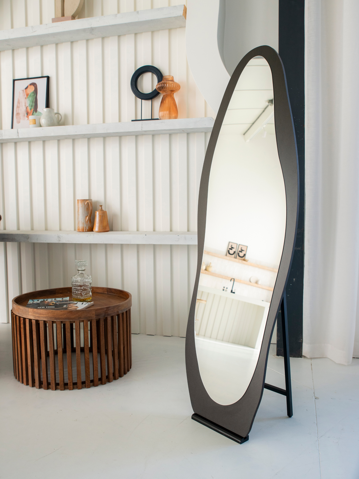 Black asymmetric full-length mirror on easel stand in styled white room beside slatted wood coffee table and wall shelves…