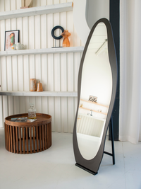 Black asymmetric full-length mirror on easel stand in styled white room beside slatted wood coffee table and wall shelves…
