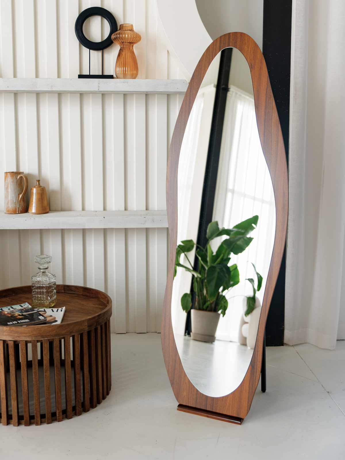 Walnut-finish asymmetric oval floor mirror leaning against white panelled wall in styled living room, reflecting potted plant