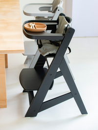 Black wooden highchair side view beside dining table, with sage green cushion, tray holding terracotta divided plate and s…