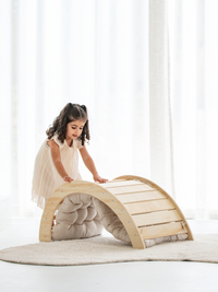 Young girl in white dress leaning over natural wood Pikler arch with beige tufted cushion underneath, bright indoor lifest…