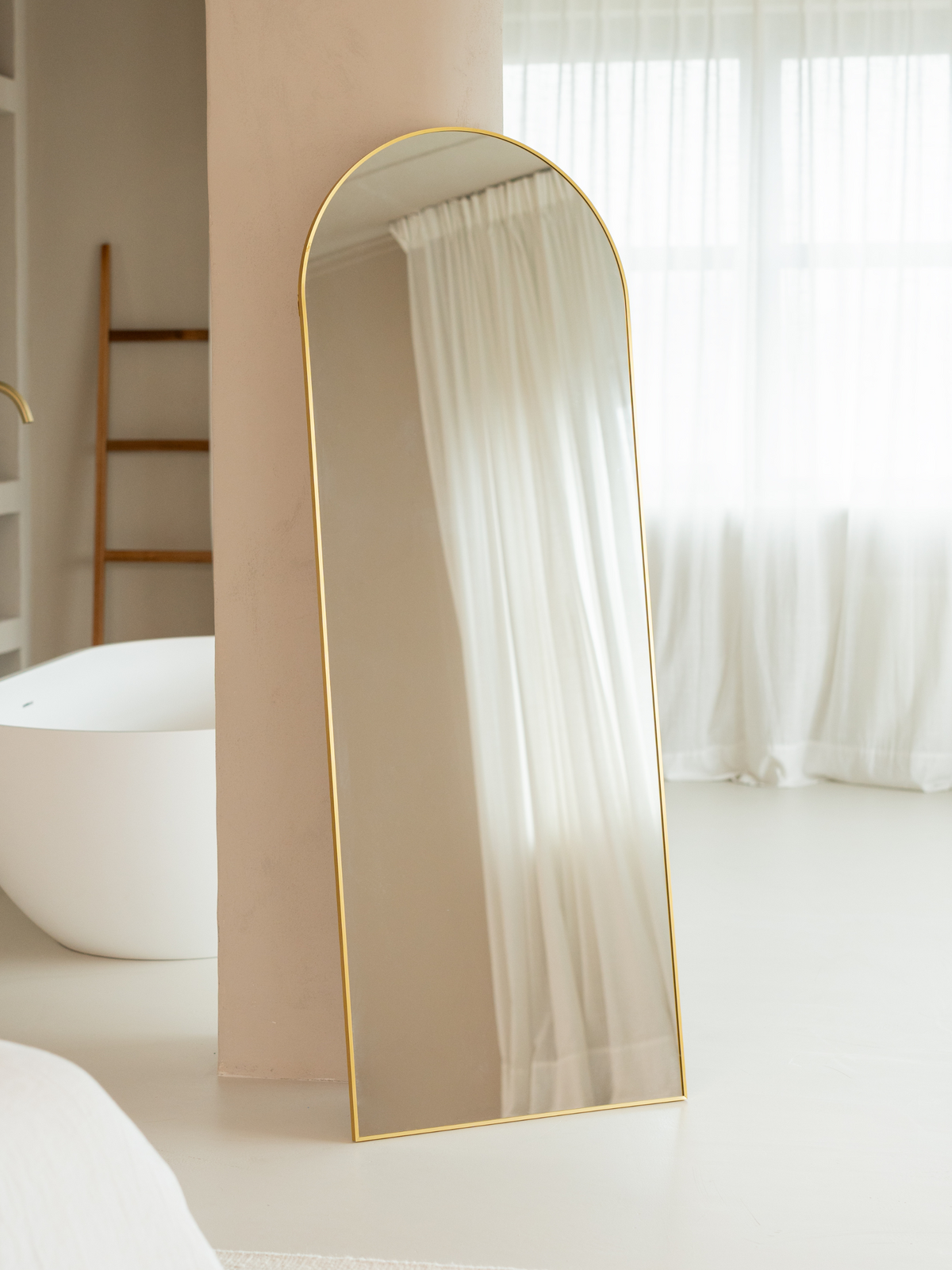 Tall arched floor mirror with slim gold frame leaning against a wall in a neutral bathroom, freestanding lifestyle view