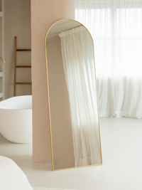Tall arched floor mirror with slim gold frame leaning against a wall in a neutral bathroom, freestanding lifestyle view