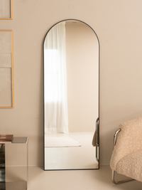 Black arched full-length mirror leaning against a beige wall in a styled room with a boucle chair and framed art nearby