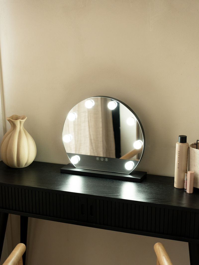 Black round Hollywood vanity mirror with glowing bulbs, on dark vanity desk beside ceramic vase and skincare products, lif…