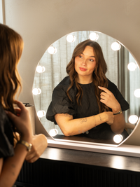 Woman sitting at vanity pointing to her hair reflected in a round white Hollywood mirror with 14 glowing bulbs, lifestyle…