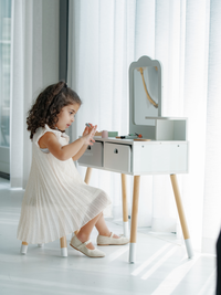 Young girl in white dress seated at white wooden children's vanity, playing with toy accessories, natural wood legs, mirro…