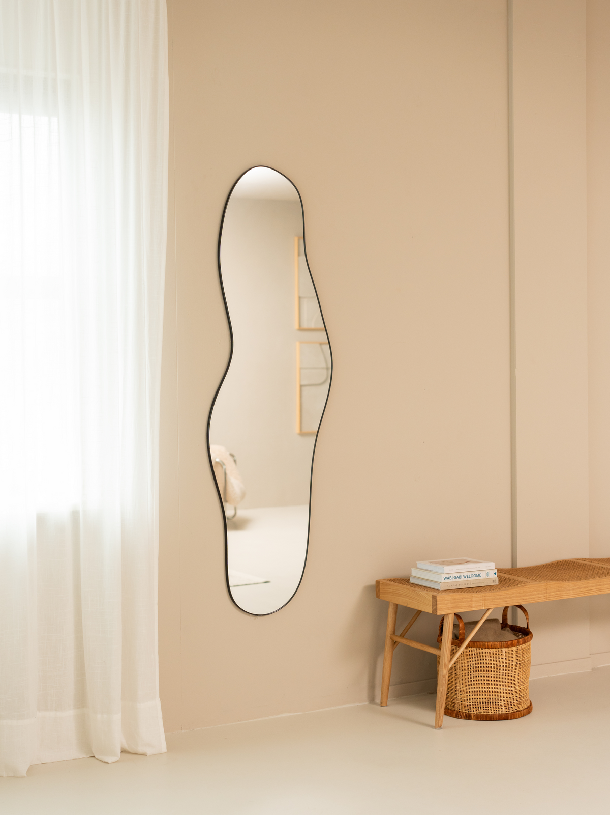 Tall wavy Julian mirror with black frame hung on beige wall, lifestyle room view with sheer curtains left and rattan bench…