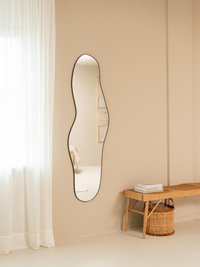 Tall wavy Julian mirror with black frame hung on beige wall, lifestyle room view with sheer curtains left and rattan bench…