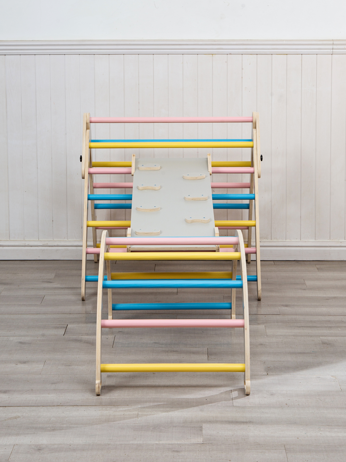 Front view of grey wooden kids climbing set with triangle frame, arch, and ramp connected, showing pink, blue, and yellow…
