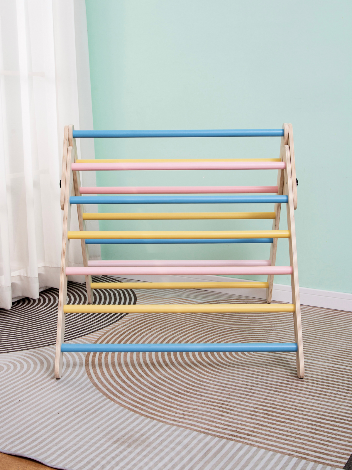 Wooden Pikler triangle climbing frame with pastel rungs, front-facing view on a striped rug against a mint wall