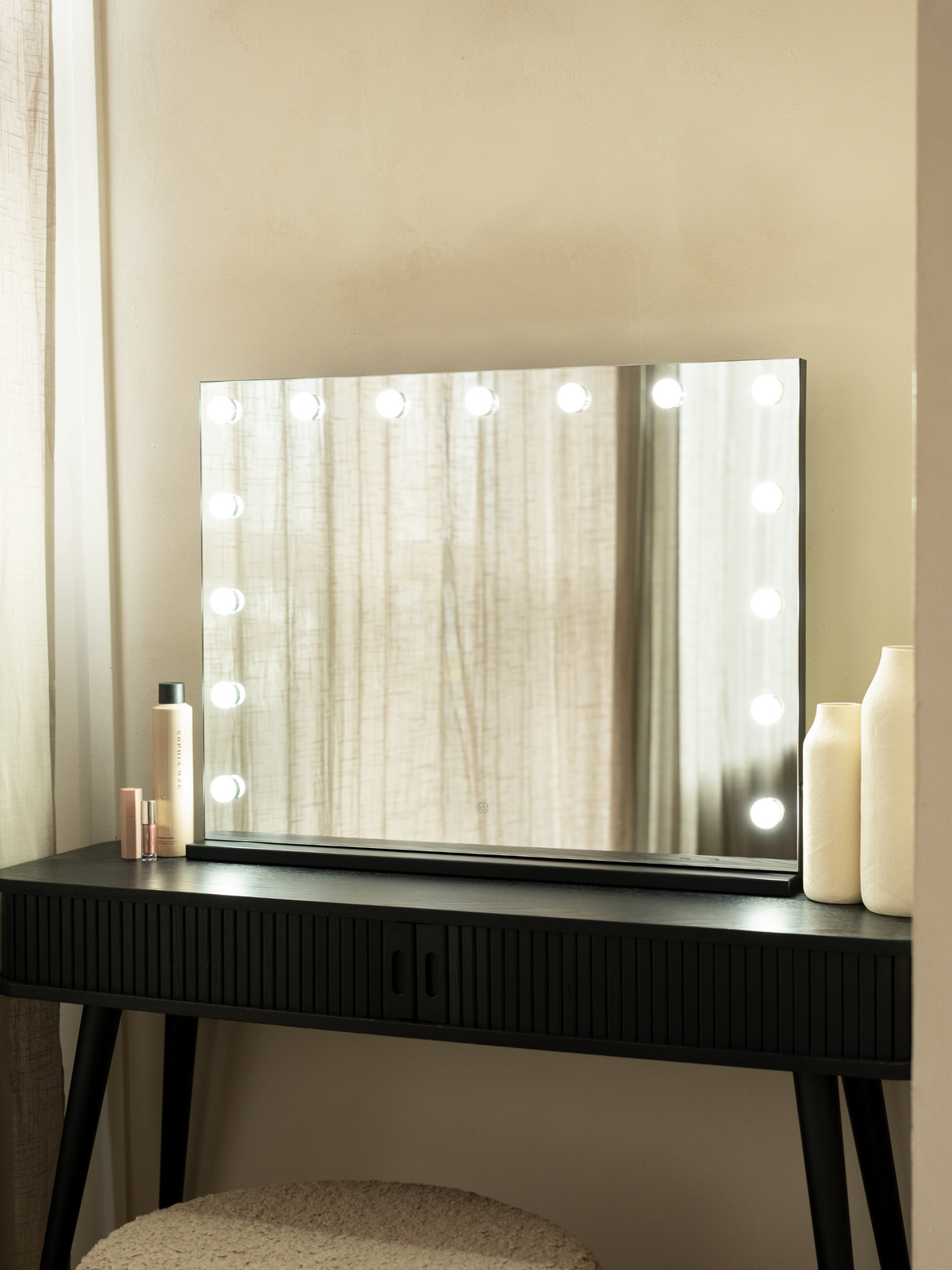 Black Hollywood vanity mirror with 15 lit LED bulbs on a dark ribbed dressing table, styled with cream vases and beauty pr…