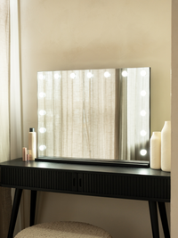Black Hollywood vanity mirror with 15 lit LED bulbs on a dark ribbed dressing table, styled with cream vases and beauty pr…
