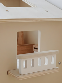 Close-up of wooden dolls house exterior showing white balcony railing with cut-out spindles beside a square window opening