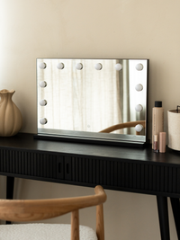 Black Hollywood vanity mirror with 12 unlit LED bulbs on dark ribbed dressing table, ceramic vase and makeup products alon…