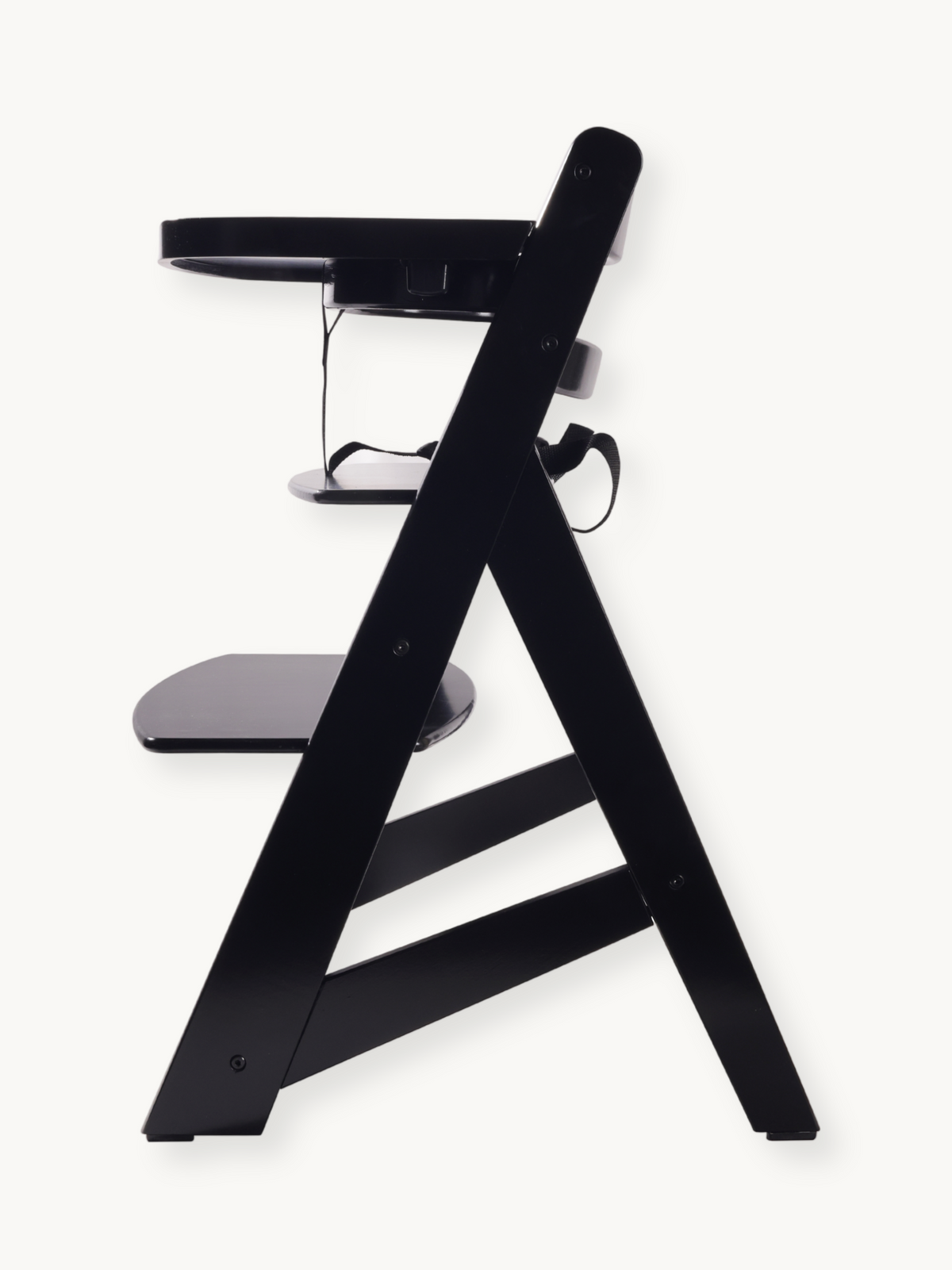 Black wooden highchair side profile, showing A-frame legs, adjustable footrest, fitted tray, and safety harness straps vis…