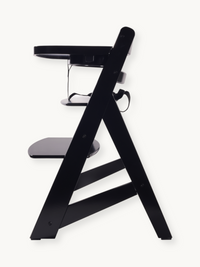 Black wooden highchair side profile, showing A-frame legs, adjustable footrest, fitted tray, and safety harness straps vis…