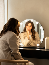 Woman seated at dark vanity table facing round black-framed Hollywood mirror with glowing surround bulbs, lifestyle bedroo…