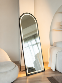 Black arched LED floor mirror on easel stand in minimal cream bedroom, warm backlight glowing, ottoman and sofa visible ne…