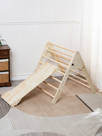 Natural wood Pikler triangle with attached slide ramp on patterned rug, angled three-quarter view in sunlit white room, no…