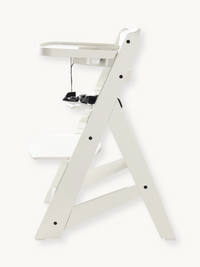 White wooden baby high chair side profile showing A-frame leg structure, attached tray, black safety harness, and black bo…