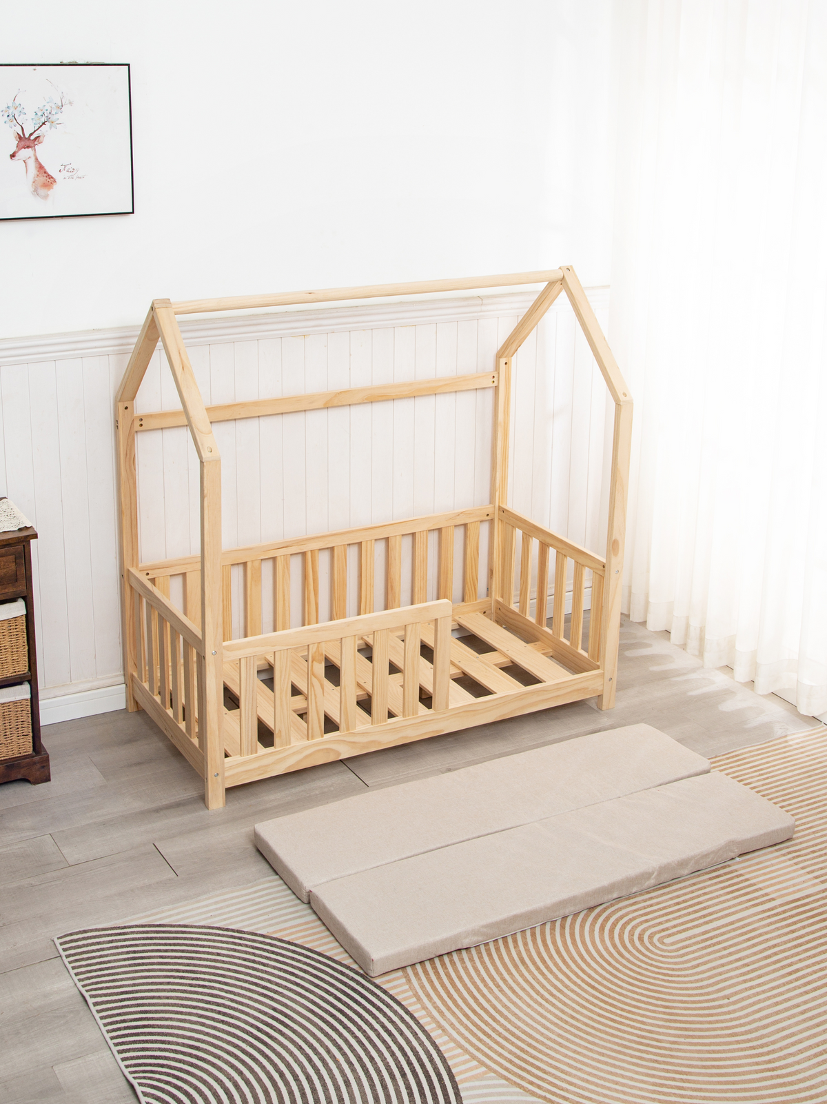 Natural pine toddler house bed with roof frame and spindle rails in styled nursery, beige folded mattress shown on rug in…
