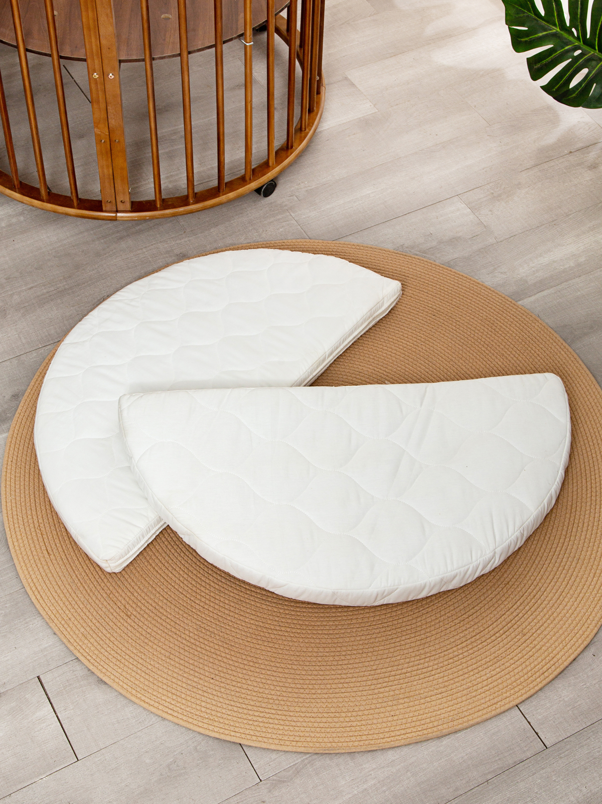 White quilted round mattress split into two half-circle sections, laid on a tan braided rug beside a round wooden baby pla…