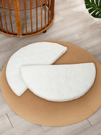 White quilted round mattress split into two half-circle sections, laid on a tan braided rug beside a round wooden baby pla…