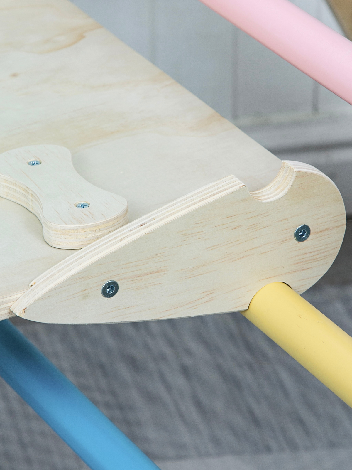 Close-up of plywood slide bracket screwed onto climbing frame rail, with pastel yellow and pink tube rungs visible