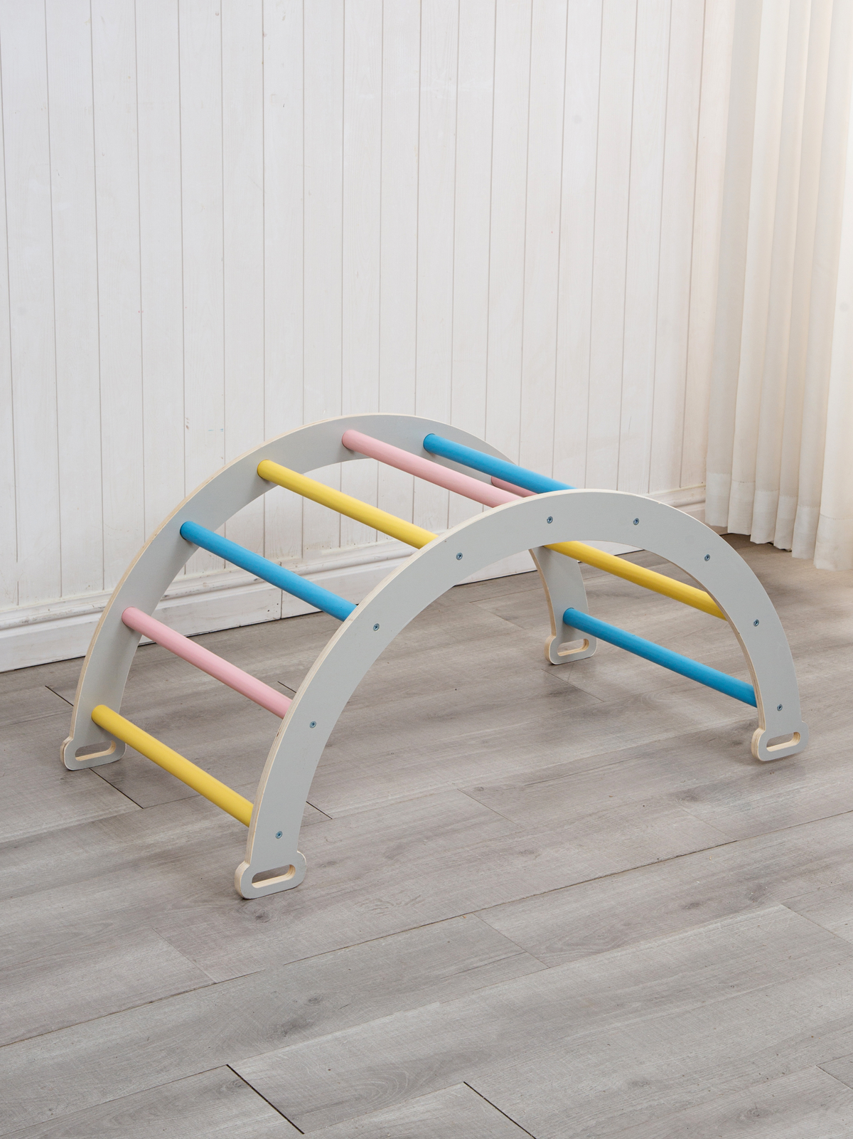 Grey wooden climbing arch with blue, pink, and yellow rungs, shown alone on light wood floor against white panelled wall