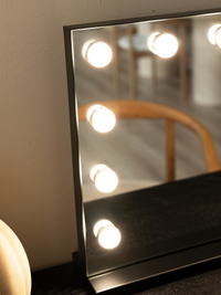 Close-up of left edge of black-framed Hollywood vanity mirror showing warm-lit LED bulbs reflected in the glass surface
