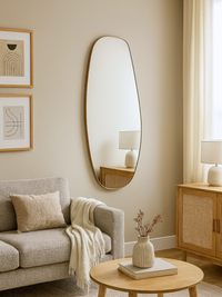 Organic-shaped gold-framed wall mirror hung in a neutral living room beside abstract art prints and a rattan sideboard