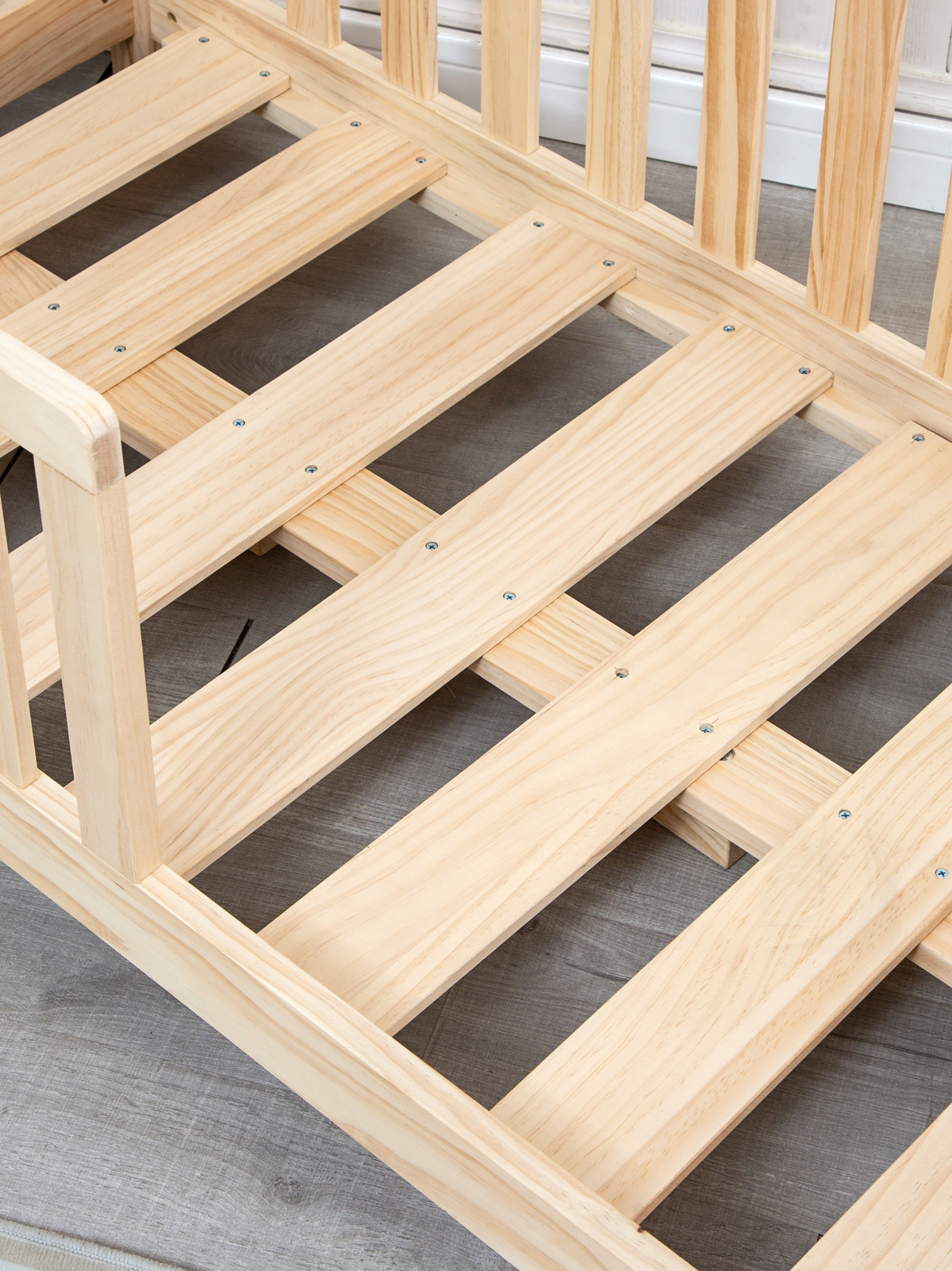 Close-up overhead view of natural pine slatted base inside assembled toddler house bed frame, screws visible, on grey floor