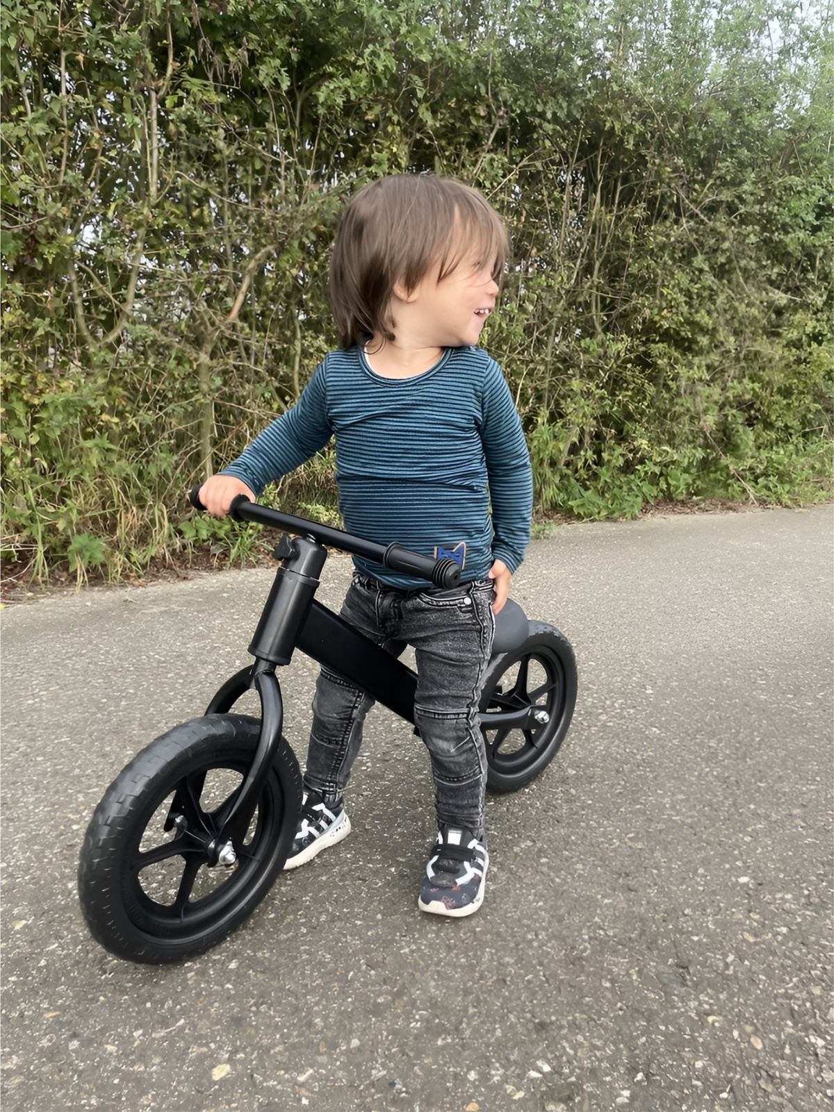 Smiling toddler straddling a matte black balance bike on an outdoor path, green hedgerow in the background