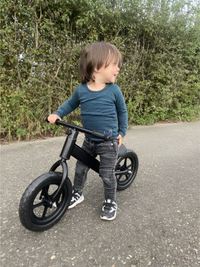 Smiling toddler straddling a matte black balance bike on an outdoor path, green hedgerow in the background