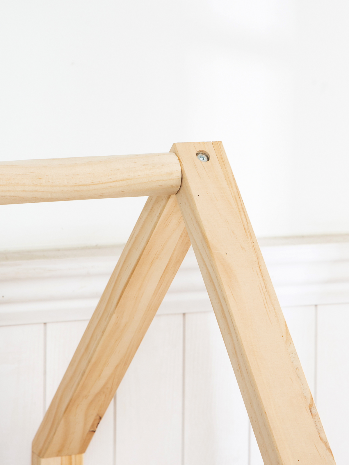 Close-up of natural pine A-frame roof joint on toddler house bed, screw hardware visible, white panelled wall background