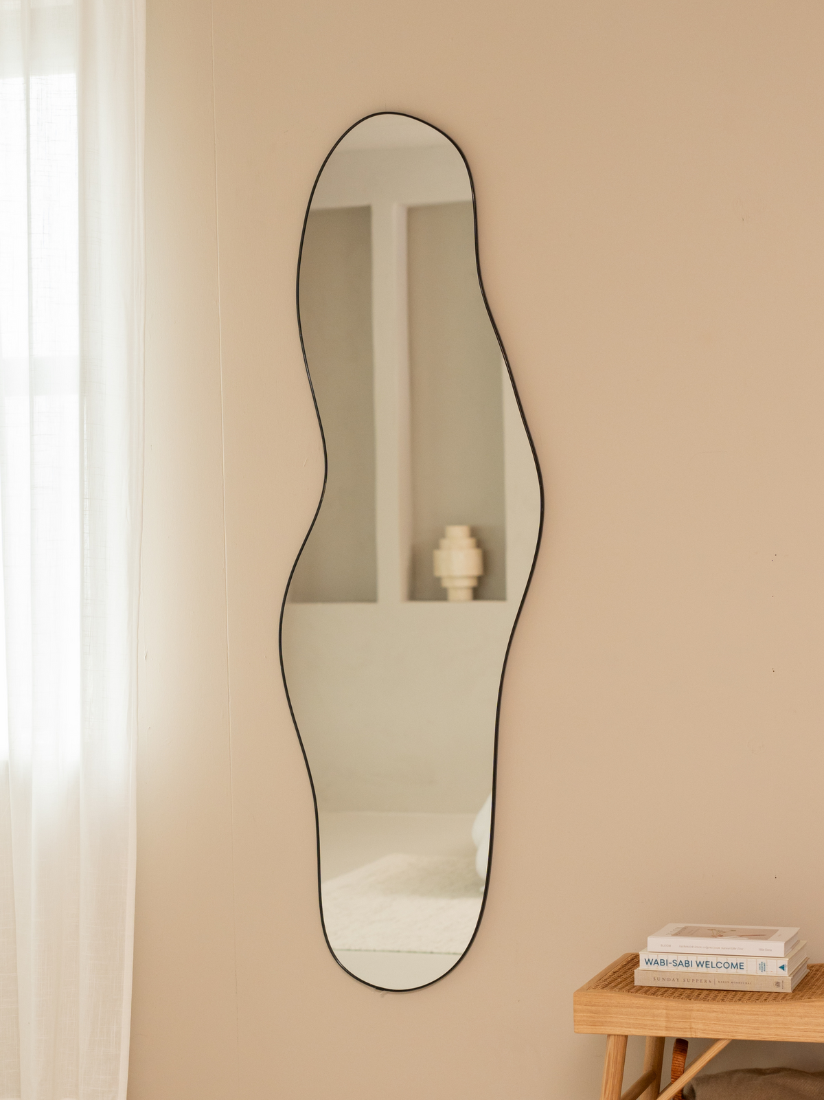 Tall asymmetric Julian mirror with black aluminium frame styled on beige wall beside rattan bench with stacked books and s…