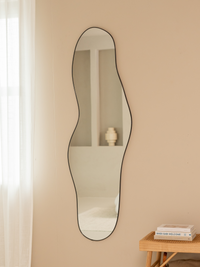 Tall asymmetric Julian mirror with black aluminium frame styled on beige wall beside rattan bench with stacked books and s…