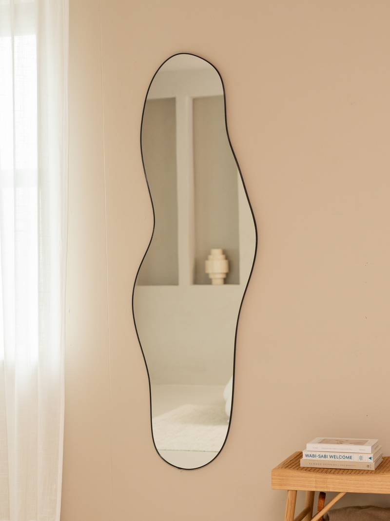 Tall asymmetric Julian mirror with black aluminium frame styled on beige wall beside rattan bench with stacked books and s…