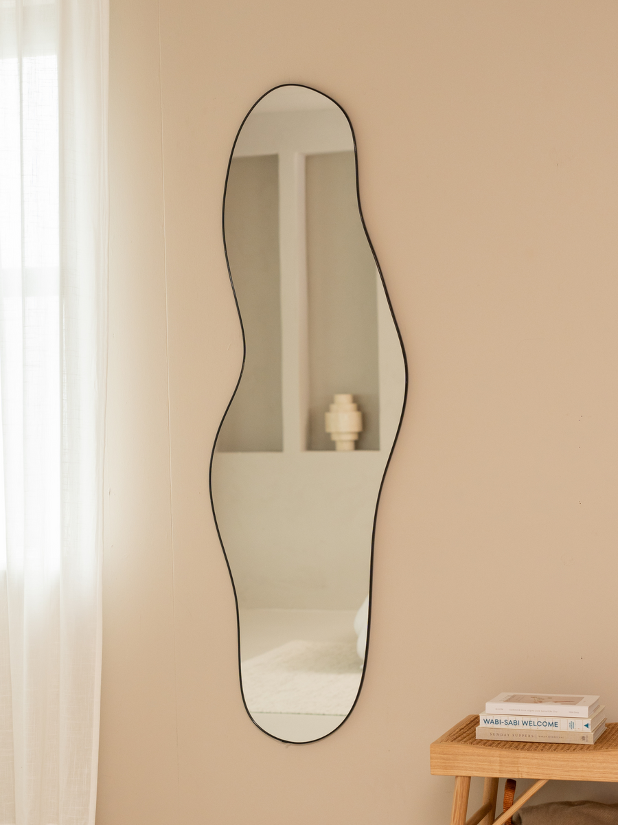Tall asymmetric Julian mirror with black aluminium frame styled on beige wall beside rattan bench with stacked books and s…